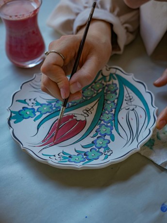 Main tenant un pinceau décorant une assiette en céramique avec des motifs floraux colorés, dans un atelier de peinture artistique.