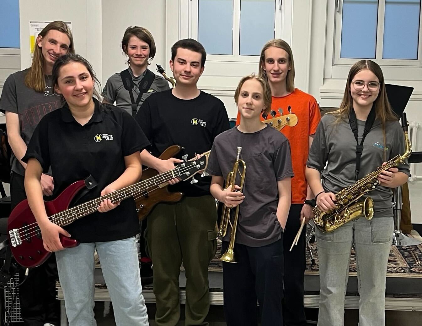 Groupe de jeunes musiciens tenant différents instruments - guitare basse, trompette, saxophone et guitare — dans une salle de pratique.