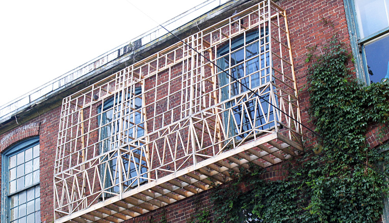 Structure en bois fixée sur la façade d’un bâtiment en briques, entourée de fenêtres et de végétation grimpante.