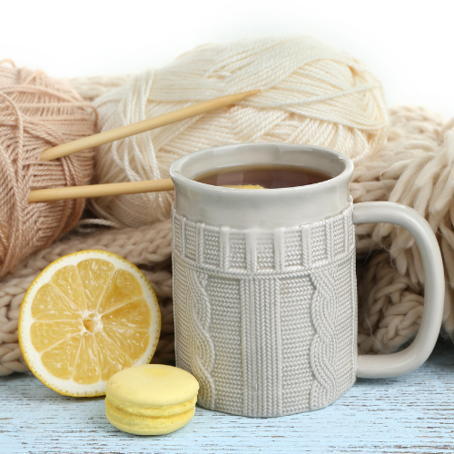 Tasse de thé posée devant des pelotes de laine et des aiguilles à tricoter, avec un citron et un macaron en avant-plan.