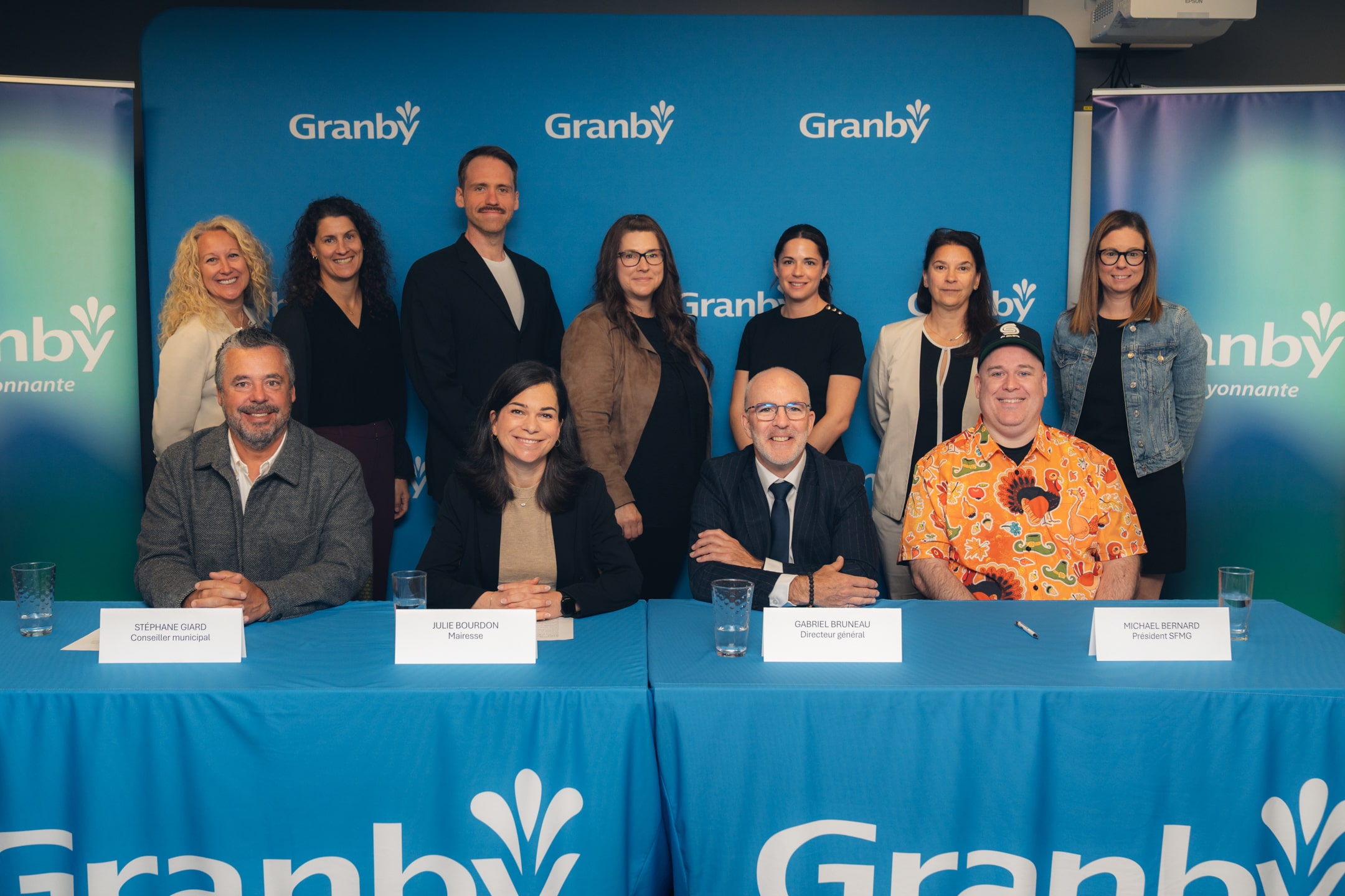 Groupe de personnes posant derrière une table avec nappe bleue “Granby”, trois personnes assises devant des cartons nominatifs. 