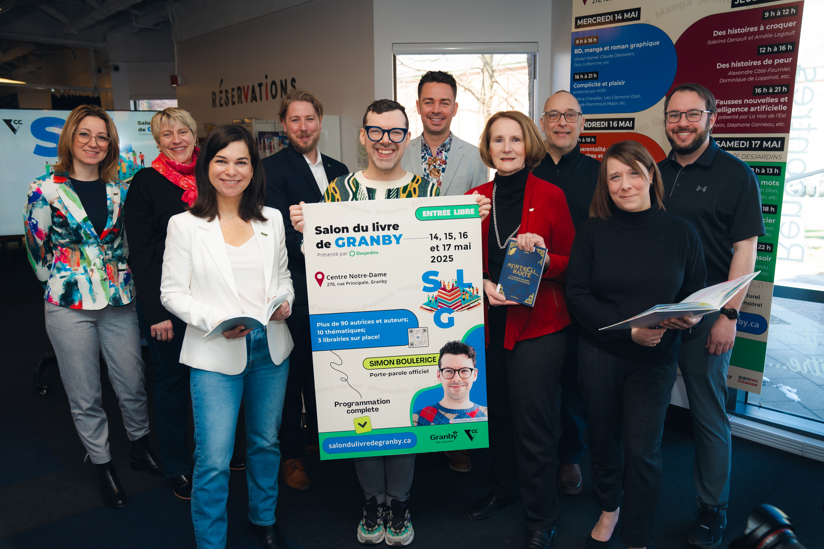 Julie Bourdon mairesse de Granby avec un groupe de personnes posant à l’intérieur d’un espace d’accueil, tenant une grande affiche annonçant le Salon du livre de Granby 2025.