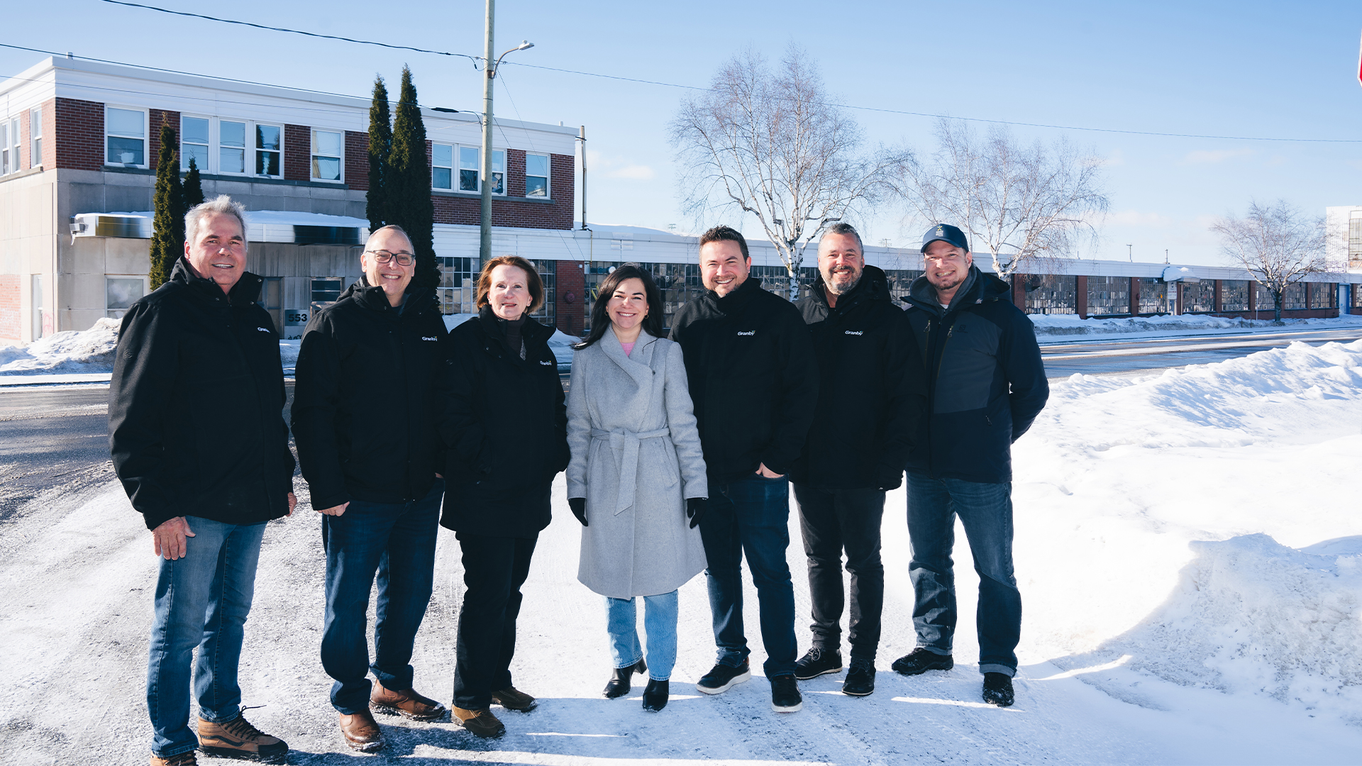 Julie Bourdon mairesse de Granby avec un groupe de personnes debout à l’extérieur sur une rue enneigée, devant un bâtiment en briques avec des arbres et un ciel clair en arrière-plan.