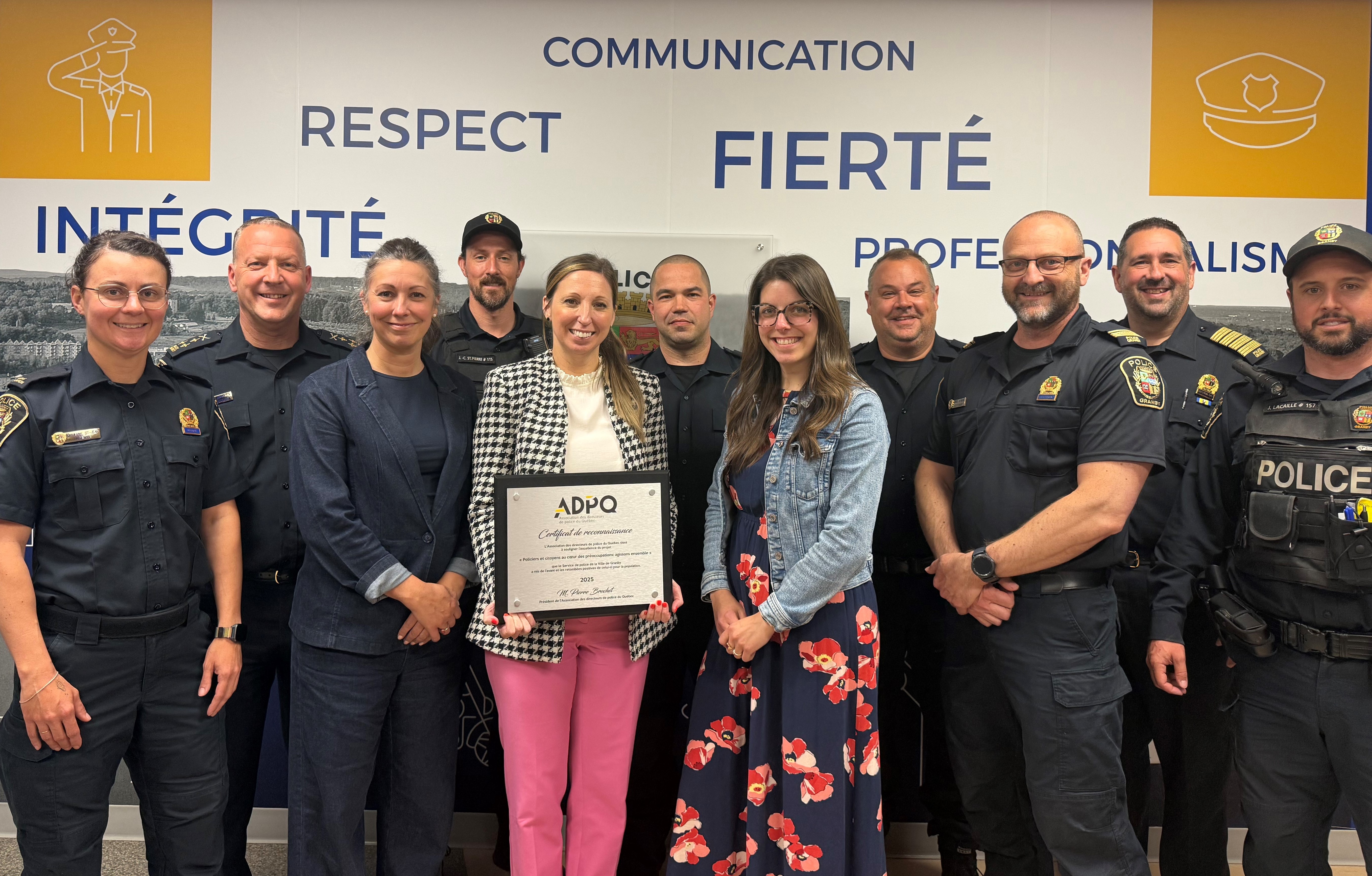 Groupe de personnes en uniforme de police et en tenue civile posant devant un mur avec des mots tels que “Communication”, “Respect”, “Fierté”, “Intégrité” et “Professionnalisme”, l’une tenant un certificat de l’ADPQ.