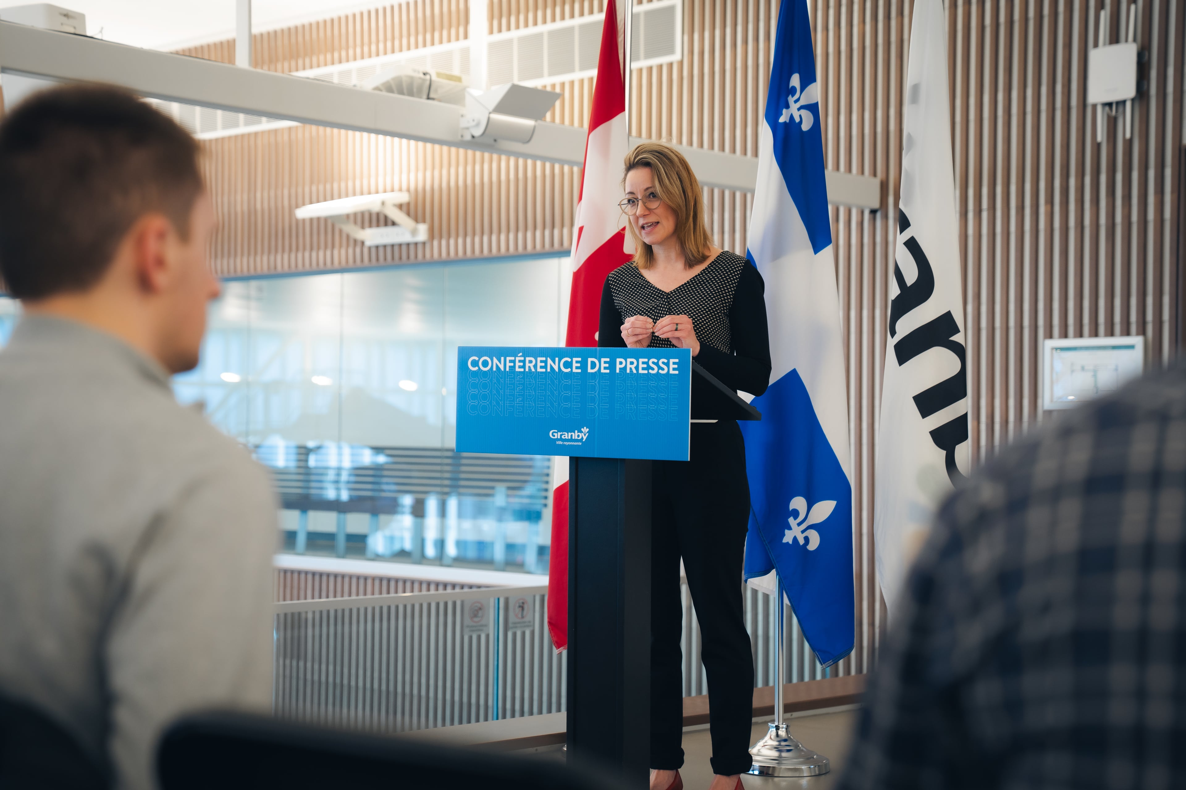 Personne debout derrière un lutrin bleu portant l’inscription “Conférence de presse” à l’intérieur d’une salle moderne. Quelques personnes sont assises en avant-plan, écoutant la présentation.