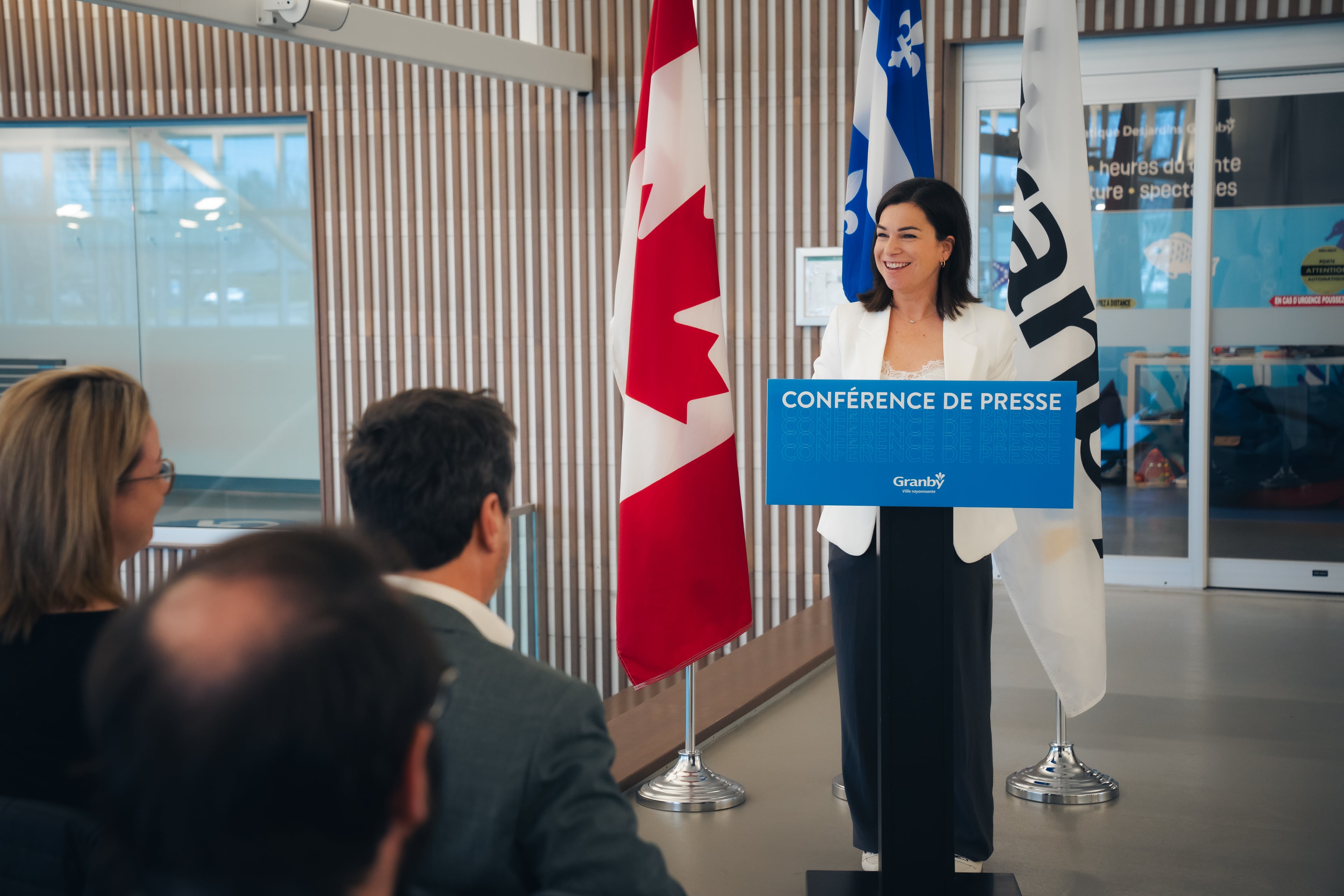 Julie Bourdon mairesse de Granby derrière un lutrin bleu portant l’inscription “Conférence de presse” à l’intérieur d’une salle moderne. Quelques personnes sont assises en avant-plan, écoutant la présentation.