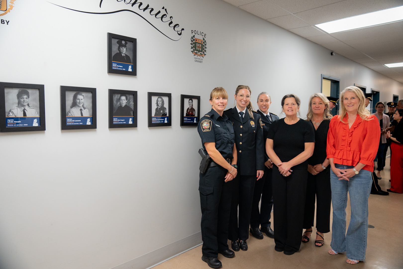 Groupe de personnes posant devant un mur décoré de cadres photo honorant des policières pionnières. 