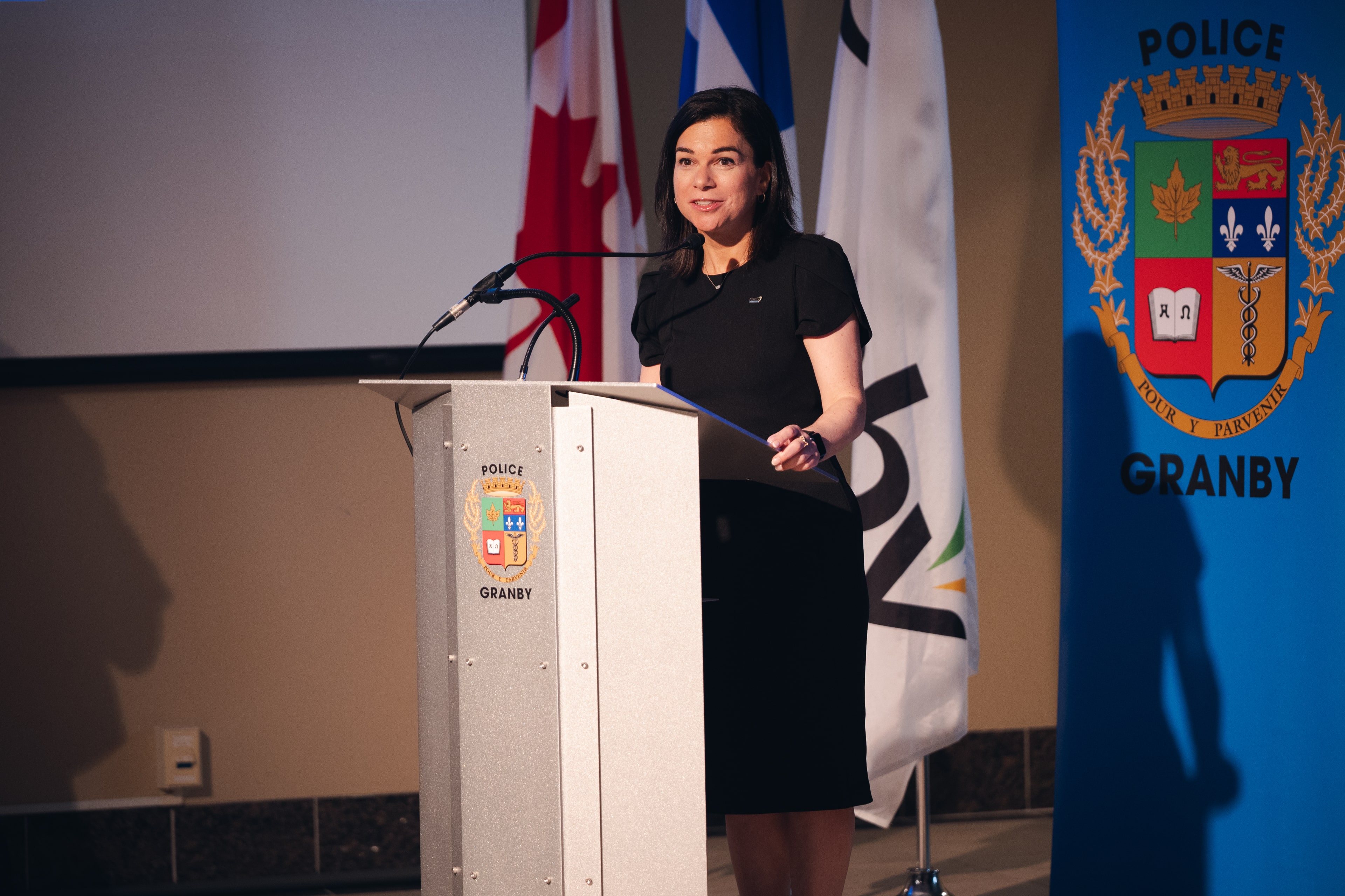 Julie Bourdon mairesse de Granby debout derrière un lutrin portant l’inscription “Police Granby”, avec les drapeaux du Canada, du Québec et de la Ville de Granby disposés en arrière-plan.