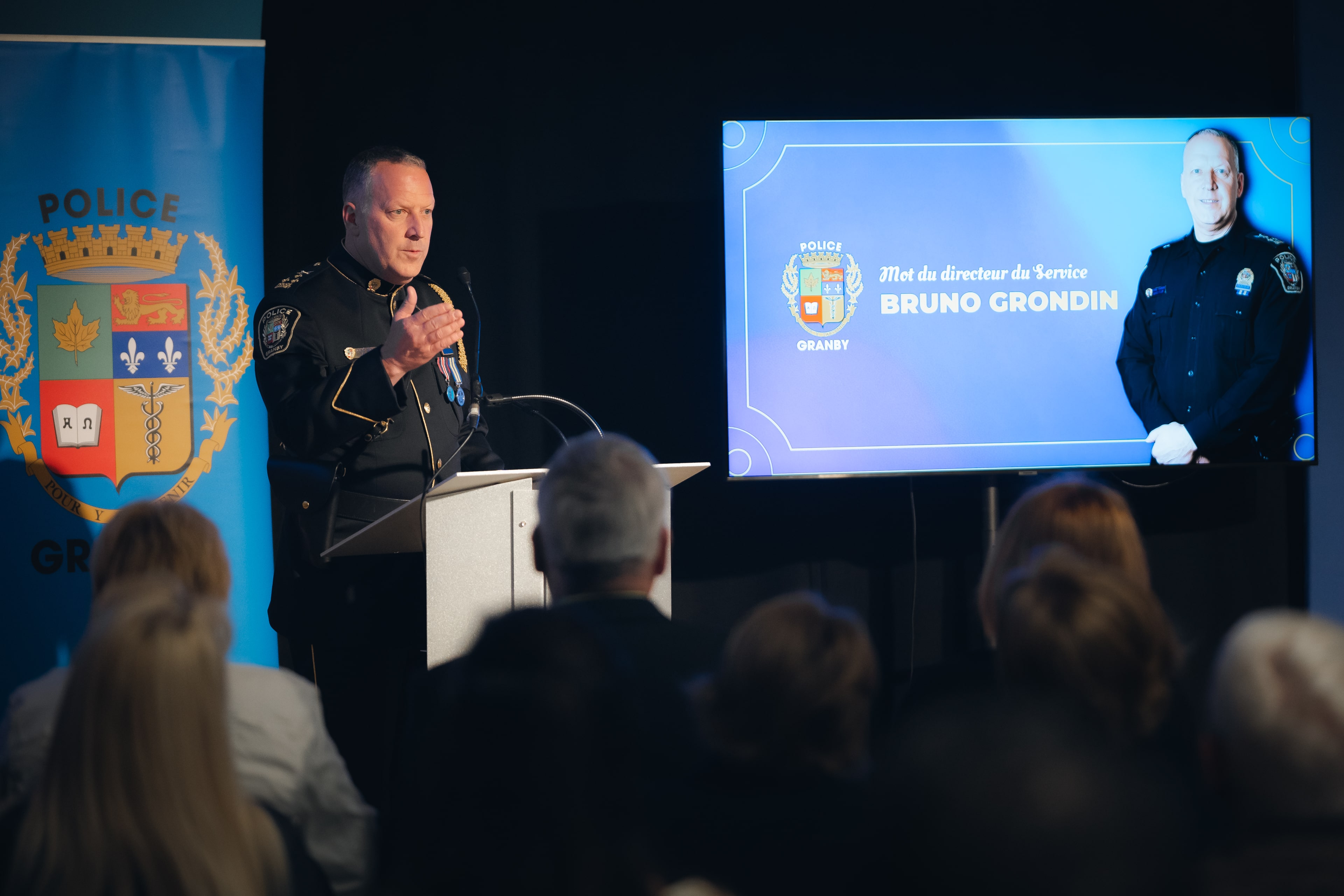 Agent en uniforme s’exprimant à un lutrin devant un public, avec un écran affichant le texte “Mot du directeur du Service Bruno Grondin” et le logo de la Police de Granby.